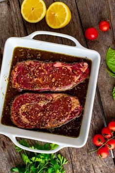 two raw meats in a baking dish with tomatoes and herbs around them on a wooden table