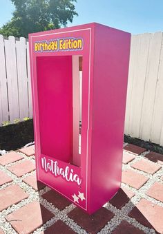 a pink birthday card vending machine sitting on top of a brick patio next to a white fence