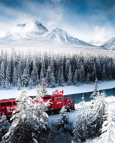 a red train traveling through a snow covered forest filled with trees and mountain range in the background