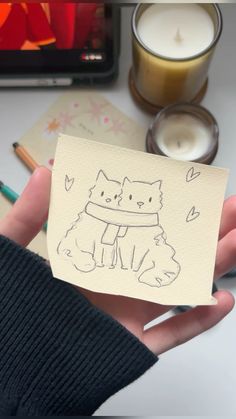 a person holding up a piece of paper with a cat drawn on it next to candles