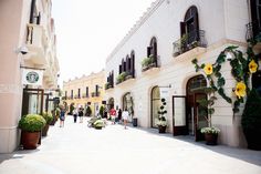 people are walking down the street in front of buildings with flowers growing on each side