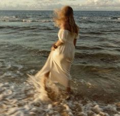 a woman in a white dress walking into the water at the beach with her hair blowing in the wind