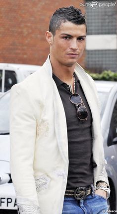 a man in a white jacket and black shirt is standing next to a car with his hands in his pockets