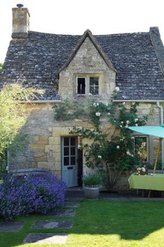 an old stone house with a table and umbrella in the front yard, surrounded by flowers