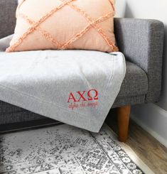 a grey couch with a red axo embroidered pillow on it's back next to a white rug