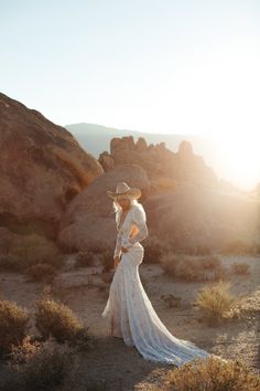 a woman in a white dress and cowboy hat standing on the side of a mountain