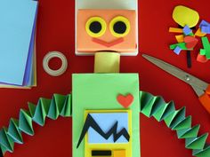 a robot made out of construction paper surrounded by other crafting supplies on a red surface