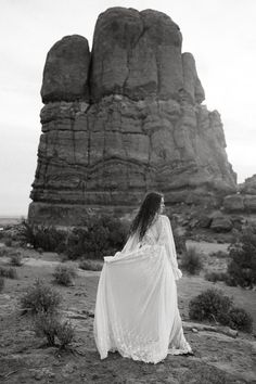 a woman in a white dress standing on top of a dirt field next to large rocks