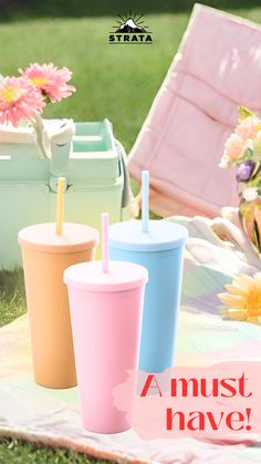 three different colored cups sitting on top of a blanket next to flowers and an umbrella