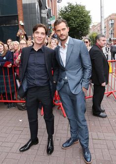 two men standing next to each other in front of a red fence with people behind them