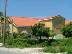 a yellow house with red roof next to palm trees and bushes on the side of the road