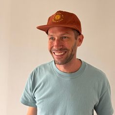 a man wearing a brown hat and blue shirt smiling at the camera while standing in front of a white wall