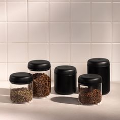 four black canisters are lined up against a white tile wall, and one is filled with coffee beans