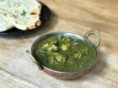 a small metal bowl filled with green sauce next to some pita bread on a wooden table