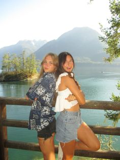 two young women standing next to each other on a wooden deck near water and mountains