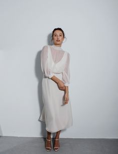 a woman standing in front of a wall wearing a white dress with sheer sleeves and heels