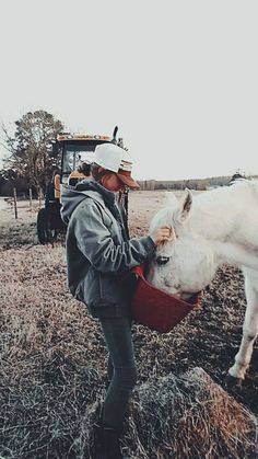 a woman is feeding a white horse some hay in a field with a tractor behind her