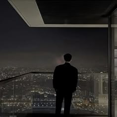 a man standing in front of a window looking out at the city lights and skyscrapers