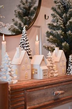 a christmas mantle with lit candles and small white houses on it, surrounded by pine trees