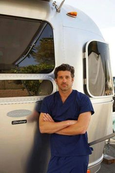 a man in scrubs leaning against the side of a camper with his arms crossed