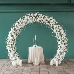 a white wedding arch with candles and flowers on the table in front of green wall