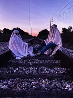 two people sitting on train tracks covered in cloths at dusk with the sun setting behind them