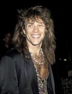 a man with long hair wearing a black jacket and leopard print shirt smiling at the camera