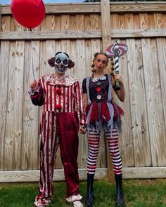 two people dressed up as clowns holding balloons and standing next to each other in front of a wooden fence