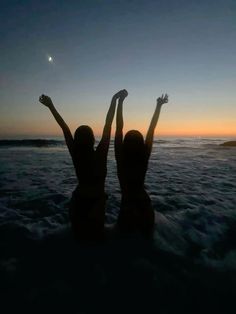 two women are standing in the water at sunset with their arms up to the sky