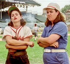 two women in baseball uniforms standing on a field with their arms crossed and looking at the camera