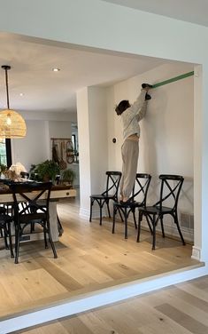 a person on a ladder painting the walls in a room with wooden floors and black chairs