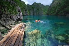 a person swimming in the water near a wooden bridge and cliffs with green trees on either side