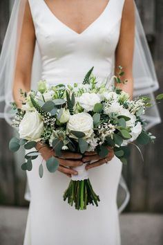 a bride holding a bouquet of white flowers and greenery on her wedding day in front of a wooden fence