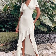 a woman wearing a white dress standing in front of some plants and trees with her hands on her hips