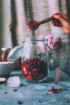 a person holding a wooden spoon over a jar filled with flowers and berries next to ice cubes