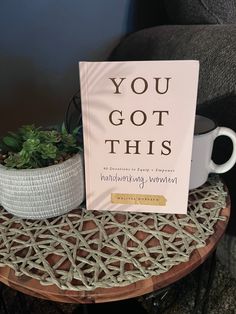 a book sitting on top of a wooden table next to a cup and potted plant