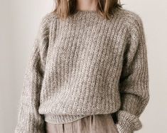 a woman standing in front of a white wall wearing a gray sweater and tan pants