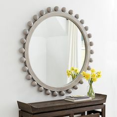 a round mirror on the wall above a wooden bench with flowers in vase next to it