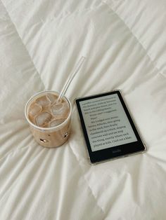 a kindle on a bed next to a cup of ice tea and a book