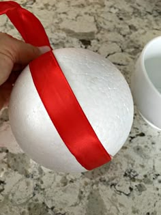 a hand holding a white ball with a red ribbon on it next to a cup