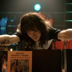 a man with long hair sitting at a table in front of a book and holding his arms out