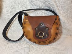 "This leather bag is made from a 1970's pattern that we dug up, it has a cool retro feel but also works well for steampunk or modern style! It is made of sienna brown bull hide, a goldish tan cowhide, and dark brown cowhide front detail with black latigo for the strap.  It is completely hand stitched with our own very strong leather cording that we make, so this is a very sturdy bag.  This one has a decorative piece on the front flap with antiqued rivets and a slot which has a brass ring that passes through to hold it closed.  The bag is 9\" wide at the widest point, 8\" tall, and the bottom is about 3.5\" deep. The strap is 2\" wide and 38\" long, can be worn on the shoulder or across the body." Brown Crossbody Shoulder Bag With Brass Hardware, Handmade Retro Brown Shoulder Bag, Retro Brown Handmade Shoulder Bag, Brown Satchel Shoulder Bag With Brass Hardware, Cognac Satchel Shoulder Bag With Brass Hardware, Retro Brown Saddle Bag For Everyday, Brown Saddle Bag With Brass Hardware, Brown Saddle Shoulder Bag With Brass Hardware, Brown Shoulder Satchel With Brass Hardware