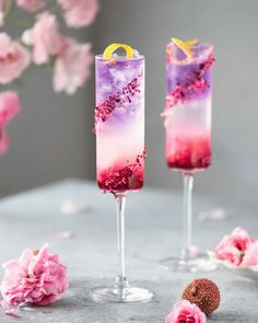 two tall glasses filled with pink and purple liquid sitting on top of a table next to flowers