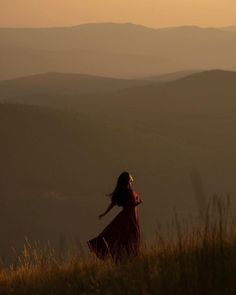 a woman in a red dress standing on top of a grass covered hill with mountains in the background