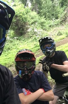 two men wearing helmets and sitting on motorcycles in the woods with their arms crossed, looking at camera