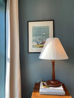 a lamp on a wooden table next to a window with curtains and a painting hanging above it