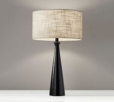 a black table lamp with a white shade on the base and a grey wall behind it