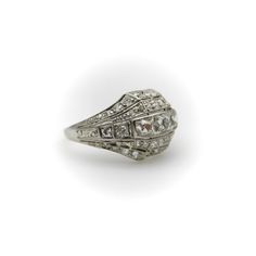 This platinum diamond ring is on the cusp of the Edwardian and Art Deco eras. The centerpiece of the ornate ring are three Peruzzi cut bezel set diamonds —one is an elongated rectangular shape, while the diamonds on either side are truncated, cut specifically for this ring. The larger diamonds are surrounded by a selection of 40 single and Old Mine Cut diamonds that are bead set into the ring. The ring has layers divided by milgrain work that tie together the lace-like patterns of the Edwardian era and the geometric designs of the Art Deco era in alternating bands. There is an added detail of a hand engraved scrolling foliate on each side of the ring. The ring has open work on the bottom that lets in the light and also reveals the triangular bottom shape of the diamonds—a nice touch that h Luxury Rings With Diamond Accents And Rectangular Stone, Luxury Rings With Rectangular Diamond Accents, Luxury Wedding Rings With Rectangular Stone, Luxury Rectangular Stone Wedding Ring, Formal Art Deco Square Cut Jewelry, Art Deco Platinum Diamond Ring With Rose Cut Diamonds, Art Deco Rings With Rectangular Stone For Formal Occasions, Heirloom Platinum Diamond Ring With Baguette Cut, Art Deco Baguette Cut Diamond Ring