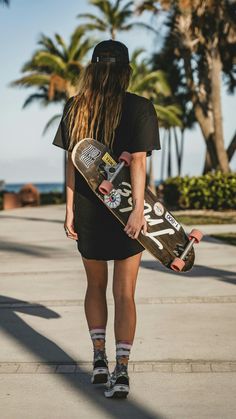 a person with long hair holding a skateboard in their hand and walking down the sidewalk