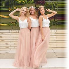 three women standing next to each other wearing long pink skirts and white tops with flowers on the wall behind them